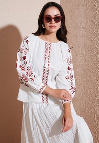 Blouse blanche avec broderie florale rouge et couture décorative, présentant de longues manches bouffantes et un col rond. Tissu élégant et léger.