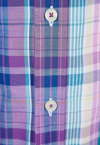 Camisa abotonada con un patrón de cuadros en tonos púrpura, azul y verde, con un botón blanco y detalle de costura visible.