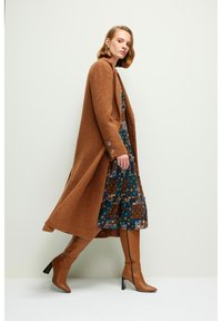 Abrigo largo de textura marrón con aberturas laterales y acentos de botones, superpuesto a un vestido de estampado floral, combinado con botas marrones hasta la rodilla.