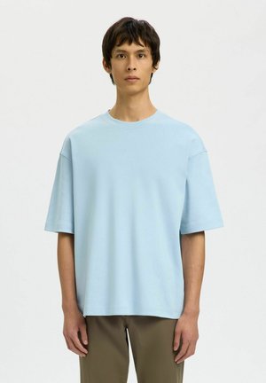 Ung mann som står vendt mot kameraet, iført en løs, lyseblå kortermet skjorte og olivengrønne bukser mot en ensfarget bakgrunn.