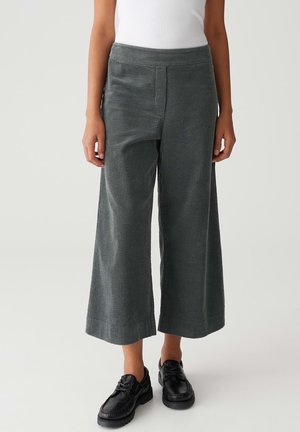 Persona vestida con pantalones de pana gris de pierna ancha y zapatos negros con cordones, de pie contra un fondo claro y liso.
