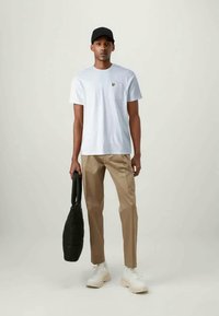 Λευκό βαμβακερό T-shirt, καφέ παντελόνι, μαύρο καπέλο και λευκά αθλητικά παπούτσια. Κρατάει μια μαύρη υφασμάτινη τσάντα με στρογγυλό σχήμα. Απλός σχεδιασμός και καθαρές γραμμές.