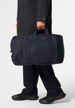 Person in dunkler Kleidung hält eine große marineblaue Reisetasche an den Griffen vor einem einfarbigen weißen Hintergrund.