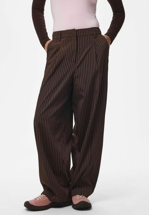 Personne portant un pantalon large à fines rayures marron foncé, des baskets roses et noires, et une chemise à manches longues avec des manches marron et un torse rose clair.