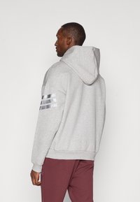 Sweatshirt cinzento feito de tecido suave, com capuz ajustável por cordão, punhos canelados e três riscas metálicas na manga esquerda.
