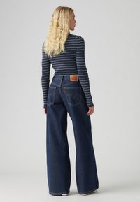 Jean bleu à jambes larges avec une taille haute, poches arrière et un patch en cuir marron de la marque. La chemise est bleu marine avec des rayures blanches, à manches longues.
