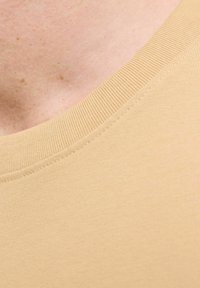 Bež majica s okruglim izrezom izrađena od glatkog i rastezljivog materijala; ima rebrasti ovratnik i suptilne detalje šivanja, prikazano u krupnom planu.