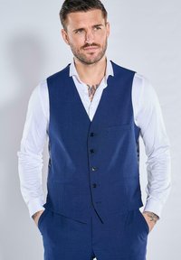 Marineblaues Weste mit strukturierter Oberfläche, ausgestattet mit einer fünf-Knopf-Verschluss, einer linken Brusttasche und Notch-Revers, über einem weißen Hemd getragen.