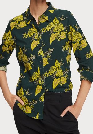 Woman wearing dark green button-up shirt with yellow floral print and rolled-up sleeves, black pants, hands in pockets.