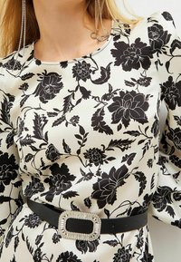 Czarny kwiatowy wzór na kremowej satynowej sukience, z dopasowanym stanem i czarnym paskiem z prostokątną, ozdobną klamrą. Długie bufiaste rękawy.