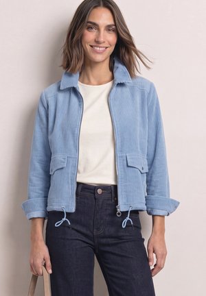 Smilende kvinde iført lyseblå ribjakke, hvid skjorte, mørke jeans, som holder en beige taskestrop mod en ensfarvet lys baggrund.