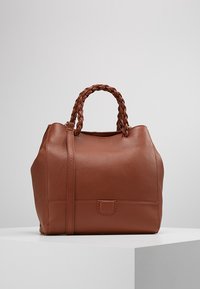 Bruine leren handtas met een gestructureerde afwerking, voorzien van gevlochten handvatten en een afneembare schouderriem. Rondvormig met subtiele details aan de voorkant.