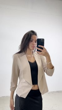 Blazer beige, entallado, con una silueta ajustada y mangas de tres cuartos, llevado sobre una blusa de encaje negra y una falda negra de cintura alta.