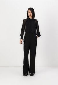 Zwarte blouse met lange mouwen en een strik halslijn, gecombineerd met zwarte broek met wijde pijpen en zwarte schoenen. De blouse heeft transparante mouwen.
