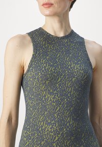Femme portant une robe sans manches à col haut avec un motif abstrait vert et gris foncé sur fond clair.