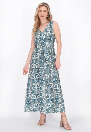Lyshåret kvinde iført ærmeløs maxikjole med V-udskæring og blå-hvidt abstrakt blomsterprint samt beige sandaler med blokhæl, poserer.