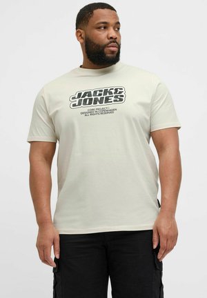 Svetlo bež majica s kratkimi rokavi z črnim grafičnim tiskom "JACK & JONES" spredaj, izdelana iz mehkega materiala, v kombinaciji s temnimi cargo hlačami.
