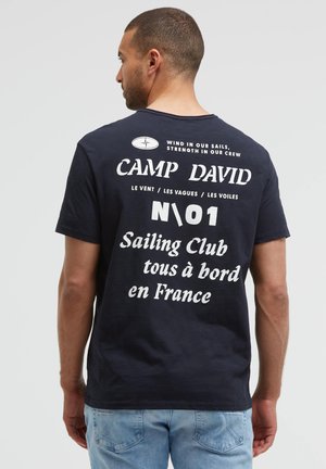Mann, der mit dem Rücken zur Kamera steht, trägt ein dunkelblaues T-Shirt mit weißem Schriftzug „sailing club“ und hellblaue Jeans vor einfarbigem Hintergrund.