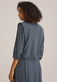 Blouse légère bleue avec un col rond, des manches bouffantes trois-quarts, une taille élastique et un léger éclat. Le dos a une ouverture en forme de trou de serrure.