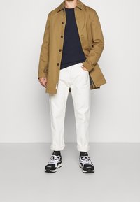 Uomo con cappotto beige, maglione blu navy, pantaloni bianchi arrotolati, sneakers bianche e nere, in piedi contro un muro chiaro e semplice.