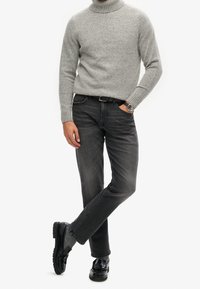 Grijze koltrui van gebreide stof, donkergrijze spijkerbroek en zwarte schoenen met een gestructureerd patroon. Horloge om de pols, model poseert casual.