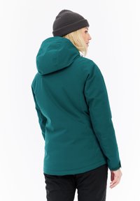 Mujer con una chaqueta con capucha en color verde azulado y un gorro de punto gris, mirando de lado con su espalda parcialmente visible contra un fondo blanco.