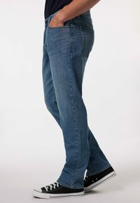 Jeans de mezclilla en un lavado azul intermedio con corte recto, que presentan bolsillos frontales y cierre de botón, combinados con zapatillas negras de tobillo bajo.