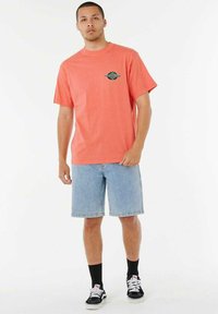 T-shirt di cotone corallo con un design grafico sul petto, abbinata a pantaloni shorts in denim e sneakers nere con accenti bianchi e calzini.