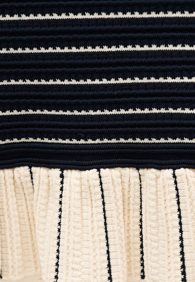 Nahaufnahme eines gerippten schwarz-cremefarbenen Strickstoffs mit horizontalen Streifen und einem gerafften cremefarbenen Abschnitt mit vertikalen schwarzen Linien.