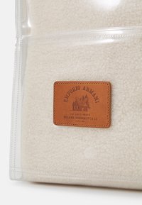 Pläd i fleece i krämfärg med en strukturerad yta, med en rektangulär brun läderetikett präglad med varumärkesdetaljer. Transparent ytterlager.