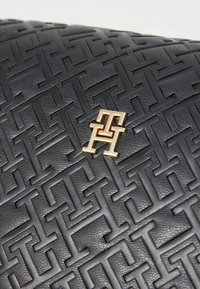 Svart lærveske med debossert geometrisk mønster, fremhevet av logo i gullfarge. Den strukturerte overflaten gir dybde.