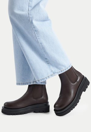 Brown leather ankle boots featuring elastic side panels, a chunky black sole, and a smooth finish. Worn with light blue wide-leg jeans.