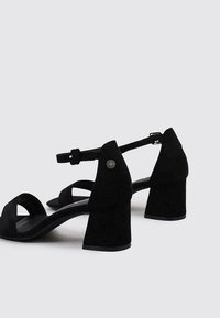 Sandalias de tacón alto de ante negro con una correa en el tobillo, punta abierta y tacón bloque. Presentan una textura suave y acentos de hardware mínimos.