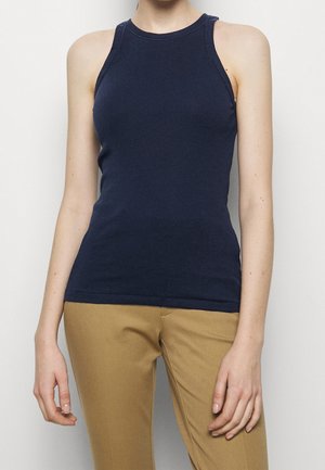 Femme portant un débardeur ajusté sans manches en bleu marine côtelé et un pantalon beige, debout devant un fond clair uni.