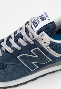 Navyblå sneakers med mocka och mesh material, med grå snörning, en stor "N"-logotyp och en vit mellansula. Klassisk design med vadderad krage.
