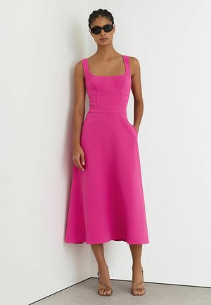 Mujer con un vestido midi sin mangas de color rosa brillante con escote cuadrado, gafas de sol negras y tacones abiertos beige, de pie contra paredes blancas.