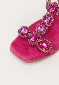 Roosa sametist sandaal, millel on nelinurkse varbaga osa, kaunistatud suurte roosade rhinestone'idega ja tekstuurse sädelevate tallaga. Komplektis on reguleeritav rihm.