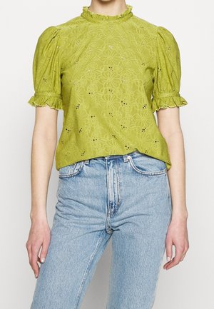 Frau trägt eine limettengrüne, mit Blumen bestickte Bluse mit gepufften kurzen Ärmeln und gerüschten Bündchen, kombiniert mit hellblauen, hoch taillierten Jeans.