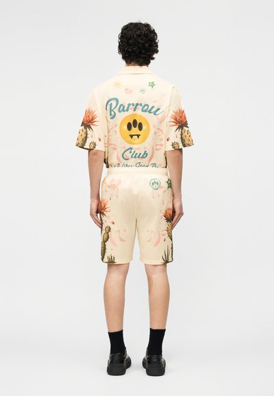 Άτομο στέκεται με την πλάτη, φοράει σετ με κρεμ πουκάμισο και σορτς με εκτυπώσεις κάκτων, ένα κίτρινο χαμογελαστό πρόσωπο με δόντια, και το κείμενο "Barrow Club".