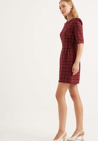 Röd rutad klänning med en figursydd design, korta ärmar och hög halsringning. Matchas med vita klackskor med blockklack. Slät textur.