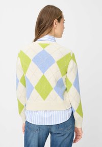 Argyle-Pullover in Creme mit grünen und blauen Rautenmuster, mit geripptem Saum und Bündchen. Darüber ein hellblau gestreiftes Hemd getragen.
