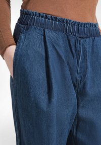 Denimbroek met een gegatherde elastische tailleband, diepe zakken en een geplooide voorkant. Donkerblauwe stof met een zachte textuur en een lichte glans.
