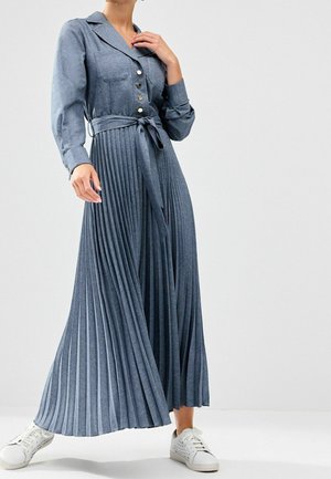 Femme portant une longue robe plissée bleue avec des boutons et une ceinture à la taille, associée à des baskets blanches, debout devant un fond uni.