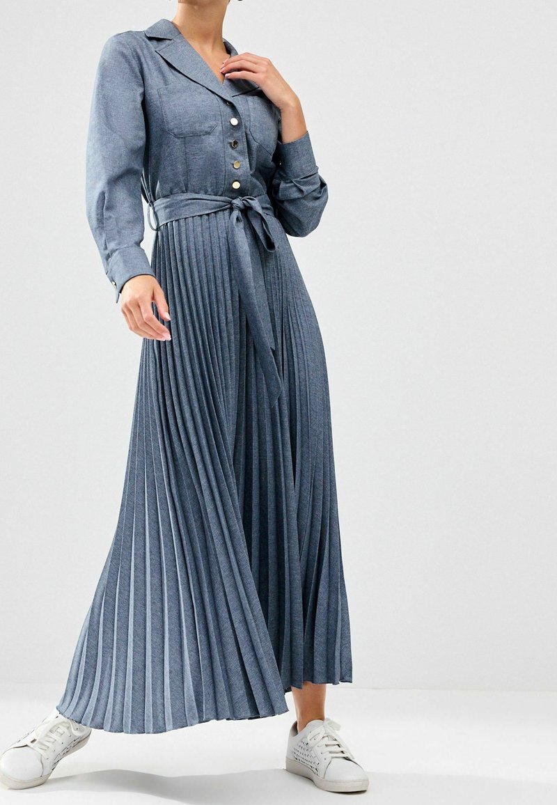 Femme portant une longue robe plissée bleue avec des boutons et une ceinture à la taille, associée à des baskets blanches, debout devant un fond uni.