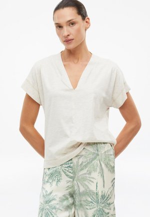 Vrouw met donker haar in een wit V-hals kortemouwtopje en lichtgekleurde korte broek met groene palmboom patronen.