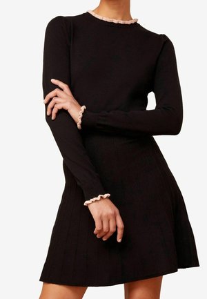 Nainen pukeutuneena mustaan, pitkähihainen mekkoon, jossa on raitainen helma ja vaaleanpunainen röyhelöreunus kaula-aukossa ja hihansuissa, seisoo käsivarret ristissä.