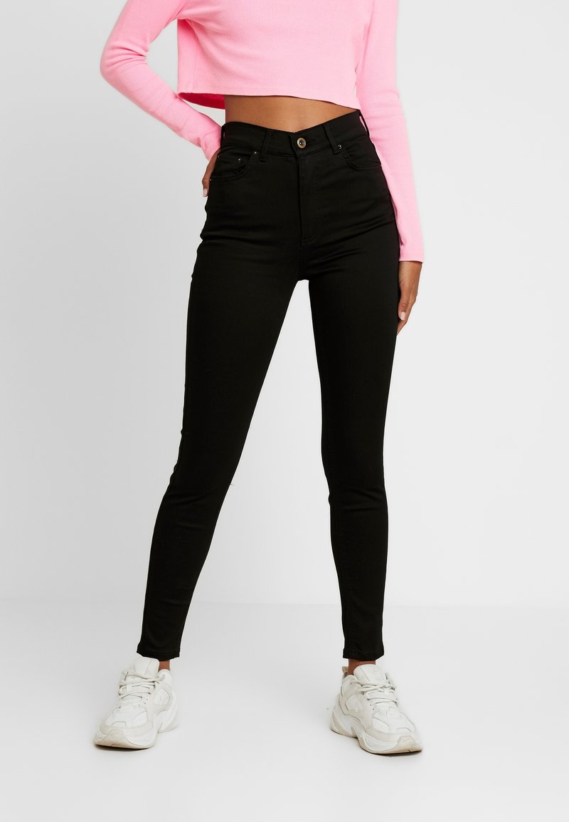 Femme en jean skinny noir taille haute, top rose à manches longues court, et baskets blanches épaisses, debout avec une main sur la hanche.
