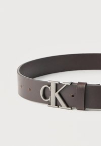 Σκούρα καφέ δερμάτινη ζώνη με αγκράφα σε σχήμα ασημένιου λογότυπου "cK" και πολλαπλές τρύπες ρύθμισης πάνω σε απλό ανοιχτό φόντο.