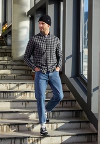 Szara i czarna koszula w kratę flanelową, niebieskie dżinsy, czarne trampki z białymi akcentami, czarna beanie, stojąc na schodach. Jasne otoczenie.