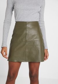 Gonna di pelle verde con un design aderente e una texture liscia. Presenta un taglio semplice e dettagli di cucitura discreti. Abbinata a un top a maniche lunghe grigio.